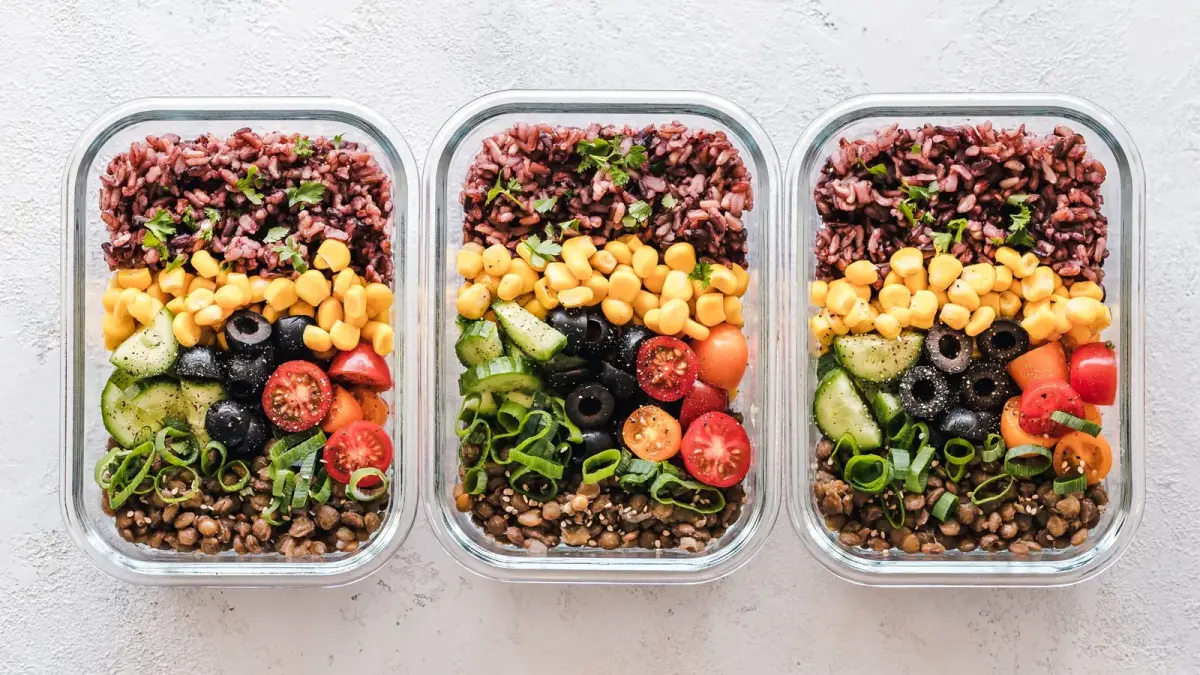 Three glass meal-prep containers filled with grains, corn, tomatoes, olives and lentils