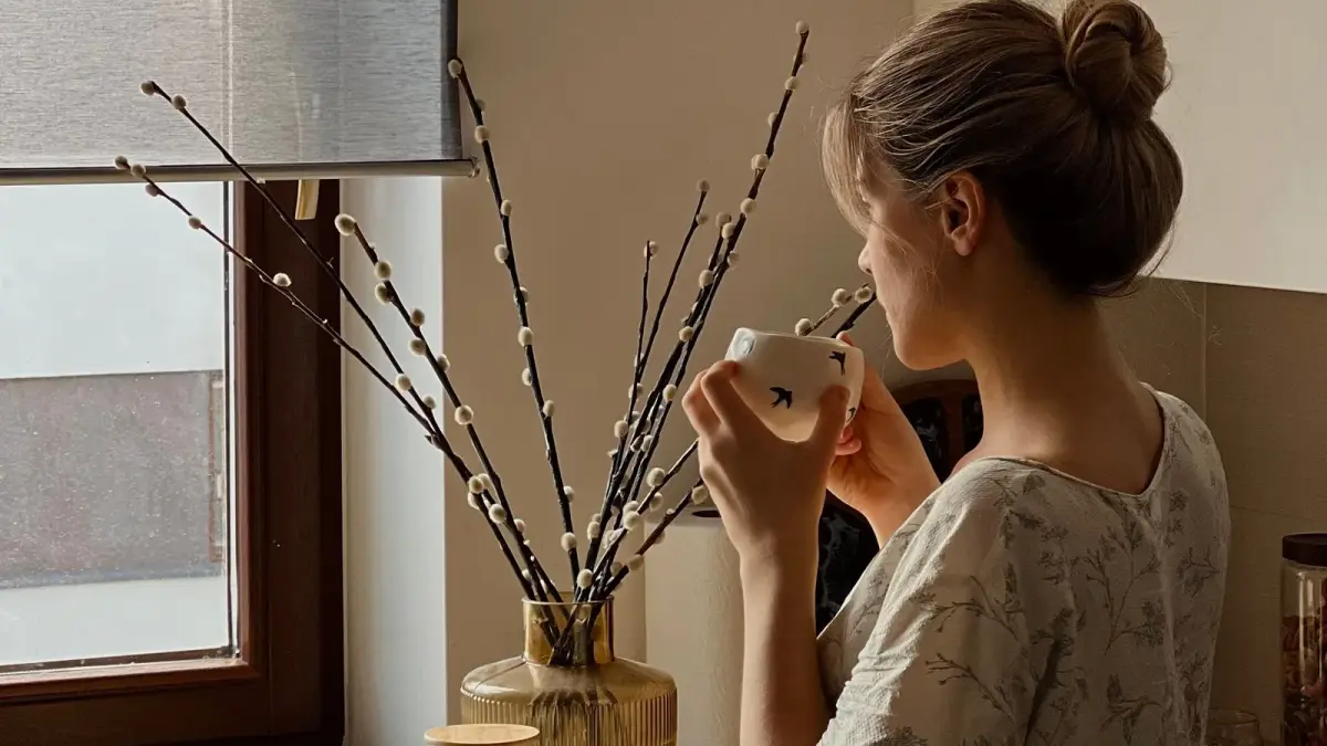 A woman drinking tea in a sunlit kitchen beside a vase of pussy willow branches